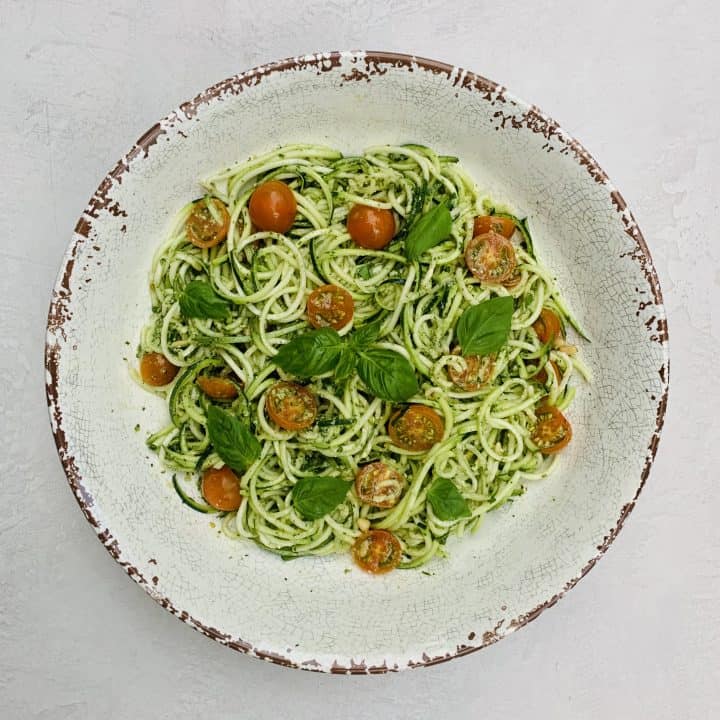 Pesto Zoodles and Tomato Salad pictured in a large serving bowl with pesto sauce in the middle and fresh basil leaves