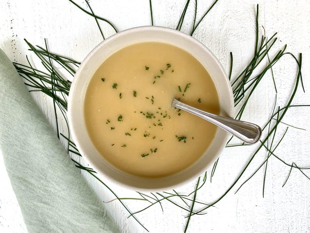 Vichyssoise Soup made with Potatoes, Cucumbers & Chives pictured in a bowl with scattered chives and a green napkin placed on a white wood board